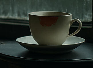 Delicate teacup with lipstick on the rim in front of a rainy window.